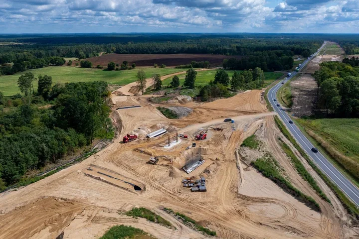 Widok z lotu ptaka na dużą placówkę budowlaną przy istniejącej drodze krajowej, gdzie prowadzone są prace ziemne i przygotowanie fundamentów pod nową infrastruk