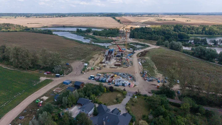 Widok z lotu ptaka na budowę mostu lub przeprawy nad rzeką na tle pól i lasów, z dźwigami oraz zapleczem technicznym na placu budowy.