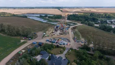 Widok z lotu ptaka na budowę mostu lub przeprawy nad rzeką na tle pól i lasów, z dźwigami oraz zapleczem technicznym na placu budowy.