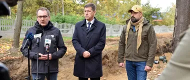 Trzech mężczyzn stoi podczas konferencji prasowej na terenie wykopalisk archeologicznych w lesie, jeden z nich przemawia do mikrofonów.