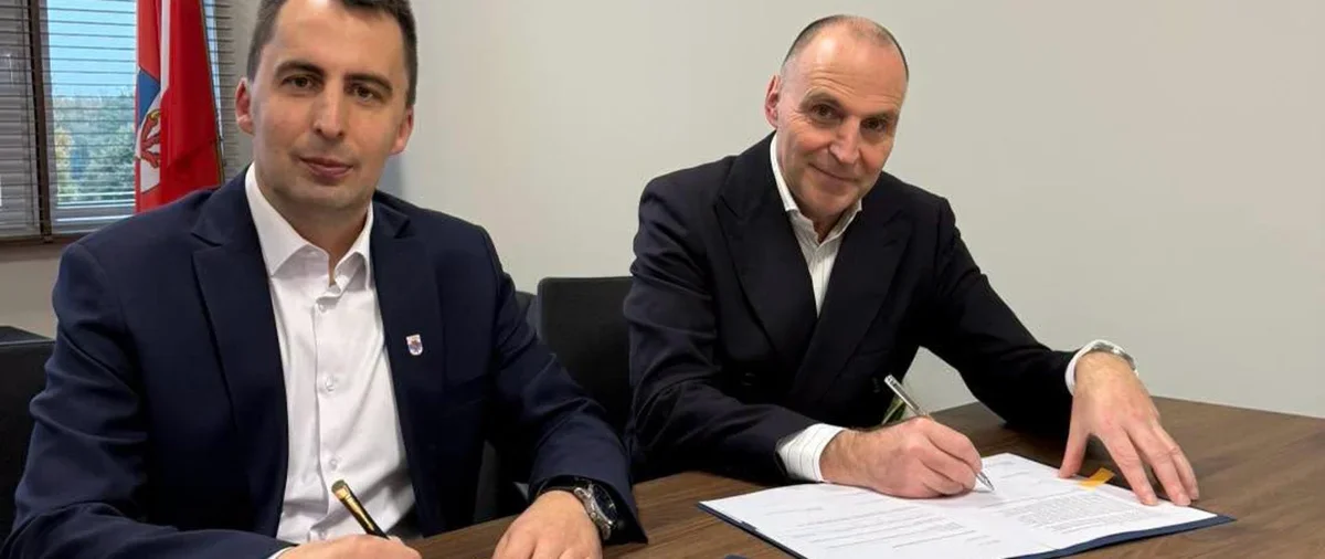 Dwaj mężczyźni w garniturach siedzą przy stole w sali konferencyjnej, jeden z nich podpisuje dokument.